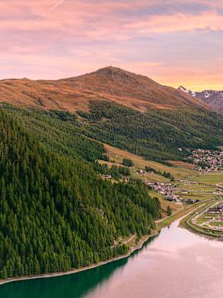 Blick über einen See auf den Ort Livigno in einem breiten Alpental bei Sonnenuntergang, umgeben von Bergen und Wäldern.