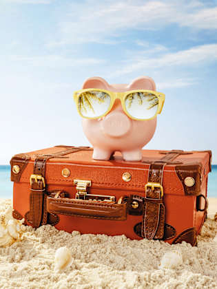 Sparschwein mit Sonnenbrille und Reisekoffer am Strand