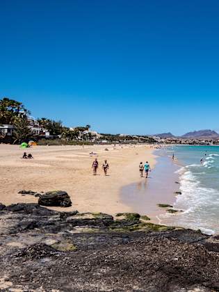Langer Sandstrand mit Spaziergängern, klares blaues Meer, an der grünen Promenade reihen sich Häuser aneinander auf Fuerteventura