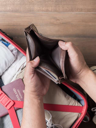 Mann hält leere Brieftasche in den Händen, darunter ein gepackter Urlaubskoffer.