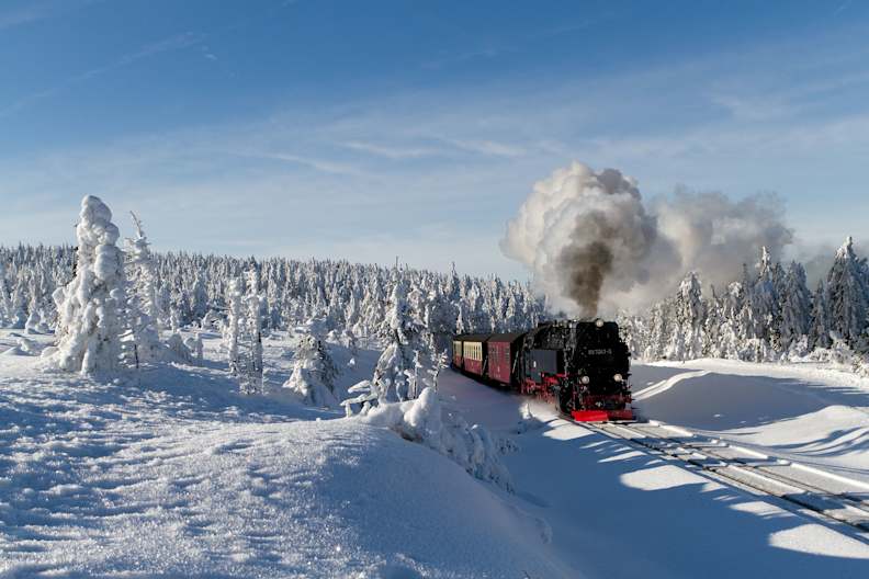 Eine schwarze Dampflok fährt durch eine tief verschneite Landschaft im Harz, umgeben von schneebedeckten Bäumen.