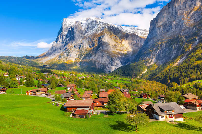 Ein kleines Dorf liegt in der sanften Hügellandschaft vor einem großen Gebirge.