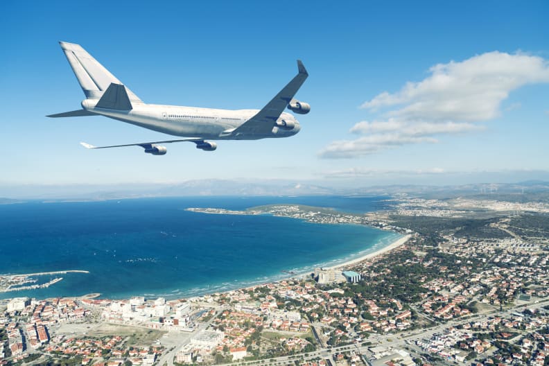 Flugzeug fliegt über eine Küstenstadt an blauem Himmel © iStock.com/jpgfactory
