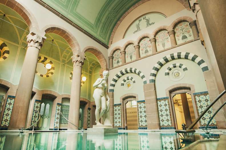Eleganter Thermalraum mit großer Marmorstatue in einem Becken, umgeben von filigran bemalten Rundbögen, hohen Säulen, Jugendstil-Ornamentik und farbigen Wandfliesen im Palais Thermal Bad Wildbad.