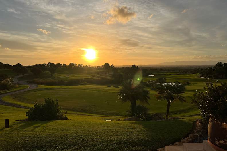 Ein weitläufiger, gepflegter Golfplatz mit Palmen und hügeligem Grün wird in warmes Licht eines Sonnenuntergangs getaucht, im Hintergrund Berge und weite Landschaft bei Palma de Mallorca.