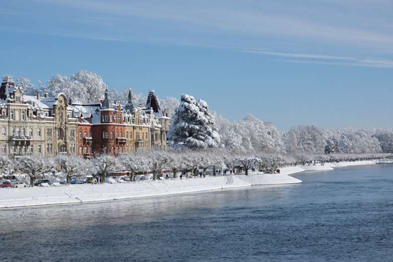 Historische Gebäude an der Uferpromenade von Konstanz, verschneite Bäume und ruhiges Wasser des Bodensees im Winter.