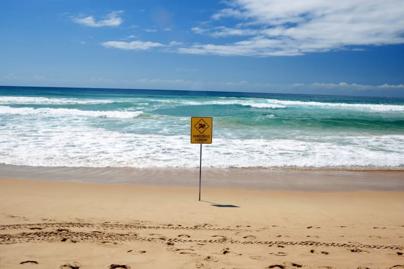Ein gelbes Warnschild an einem Strand direkt am Meer.