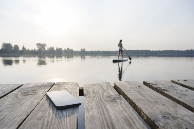 Ein Handy liegt auf einem Holzsteg, während eine Frau auf dem See Stand-Up paddelt.