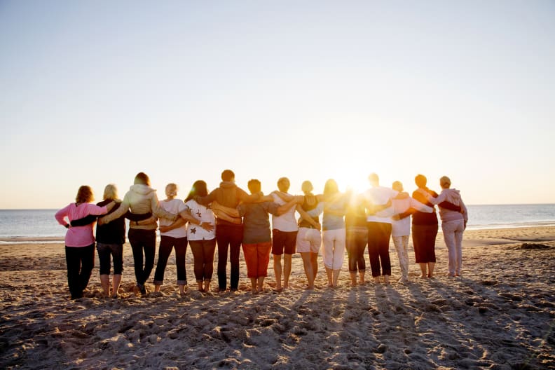 Eine Gruppe Menschen schaut mit den Armen umeinander von einem Strand aufs Meer.