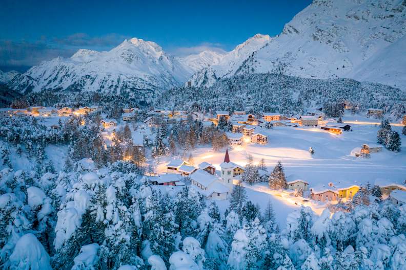 Alpendorf Maloja und Chiesa Bianca umrahmt von verschneiten Wäldern in der Abenddämmerung, Bergell, Kanton Graubünden, Engadin, Schweiz. © Roberto Moiola / Sysaworld via Getty Images