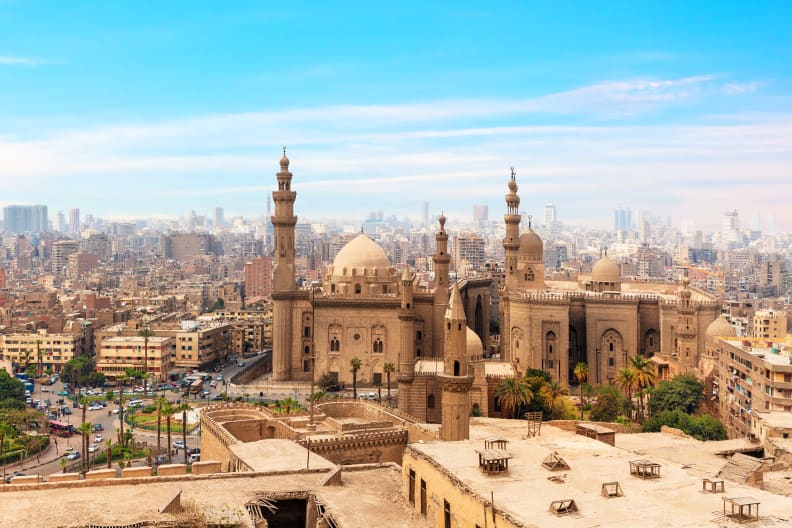 Die Moschee Madrassa ist im Vordergrund mit ihren vielen Türmen zu sehen. Im Hintergrund liegen unzählige Häuer der Stadt Kairo. In der Ferne erkennt man Pyramiden.