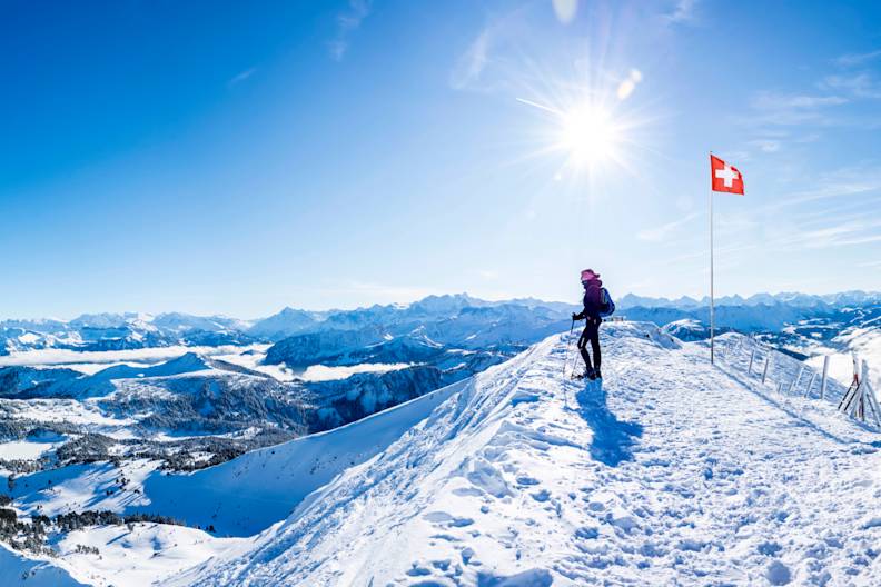 Ein Wanderer steht vor einer Schweizer Fahne auf einem Berg in den Alpen.
