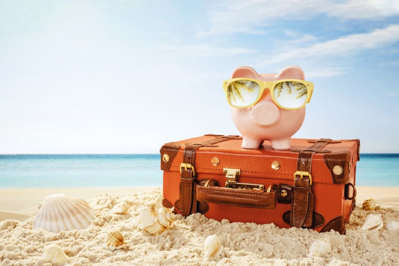 Sparschwein mit Sonnenbrille und Reisekoffer am Strand