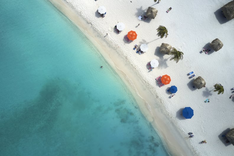 Ansicht von oben. Links ist das Meer, rechts ein Strand mit Sonnenschirmen, Palmen und Strandliegen.