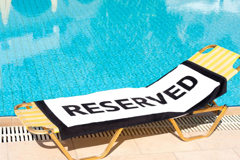 Ein Strandtuch mit dem Wort "Reserviert" auf einem Liegestuhl neben einem Swimmingpool. © clubfoto/iStock / Getty Images Plus via Getty Images