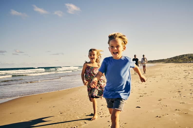 zwei kleine Kindern, die mit ihren Eltern im Hintergrund am Strand laufen © iStock.com/pixdeluxe