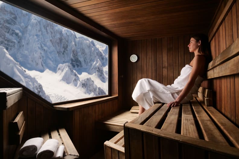 Frau sitzt in einer Sauna und blickt durch ein Fenster in schneebedeckte Berge.