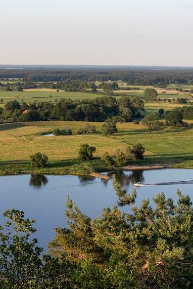 Blick über eine grüne flache Landschaft. Im Vordergrund fließt ein breiter Fluss, auf dem ein kleines Boot fährt.