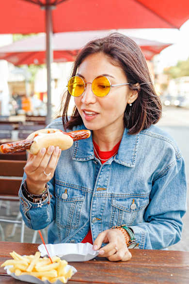 Zwei Frauen sitzen an einem Tisch unter roten Schirmen an einem Imbiss, die eine isst Currywurst mit Brötchen und die andere Bratwurst mit Pommes.