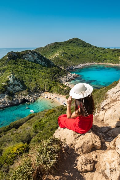 Frau mit Sonnenhut sitzt hoch oben auf einem Felsen und schaut auf Meer © iStock.com/RistoArnaudov