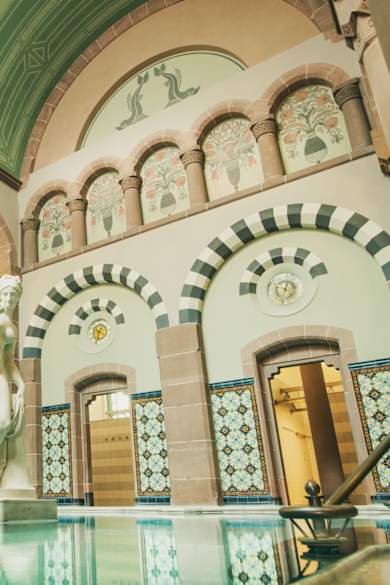 Eleganter Thermalraum mit großer Marmorstatue in einem Becken, umgeben von filigran bemalten Rundbögen, hohen Säulen, Jugendstil-Ornamentik und farbigen Wandfliesen im Palais Thermal Bad Wildbad.