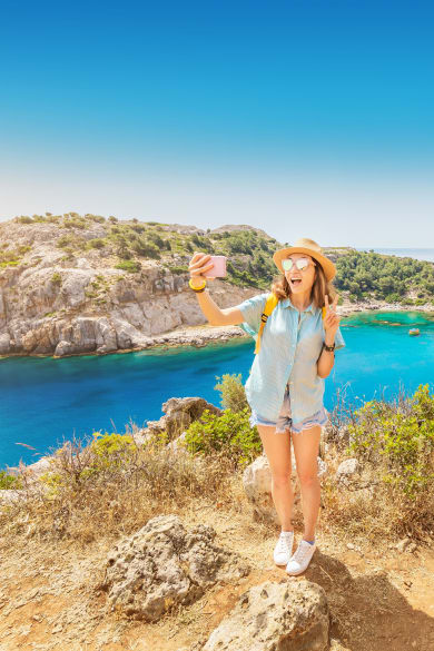 Mädchen mit Hut macht ein Selfie mit einer azurblauen Mittelmeer-Bucht im Hintergund © iStock.com/frantic00