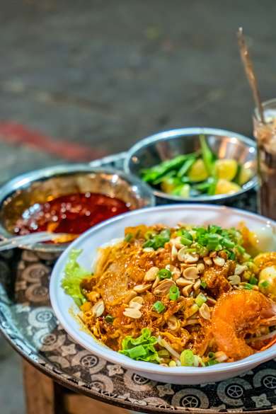 Auf einem kleinen Beistelltisch steht ein Teller vietnamesisches Mi Quảng, eine Soße und Getränke.