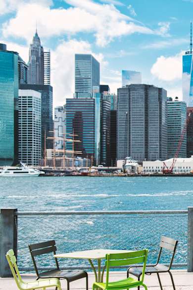 Eine Frau in einem blauen Kleid steht an einem Brückenzaun. Auf der anderen Seite liegt ein Fluss und eine Skyline.