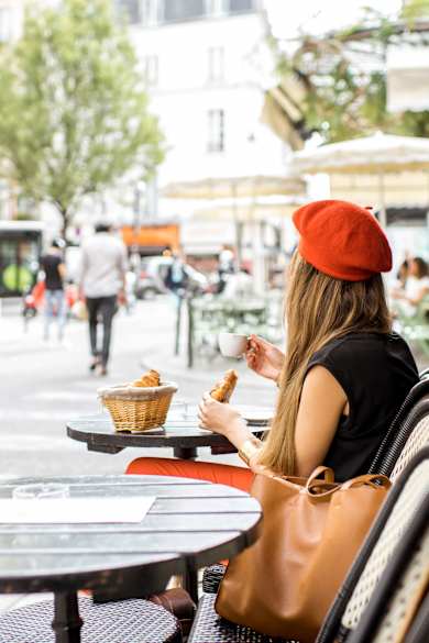 Eine Frau mit rotem Beret sitzt an einem Cafétisch und hält ein Croissant und einen Espresso in den Händen.