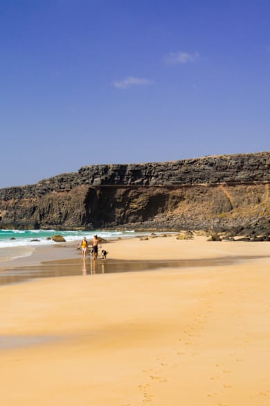 Blick auf die Naturstrand-Bucht Playa del Águila, Fuerteventura mit Steilküste und zwei Menschen mit Hund am Strand
