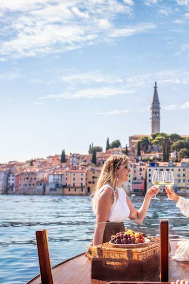 Zwei Frauen sitzen auf einem Boot auf dem Meer vor der Kulisse eines bunten Hafens in Kroatien und stoßen mit Weingläsern an