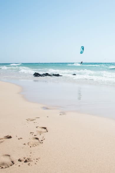 Sandstrand und blaues Meer mit Fußabdrücken und Menschen die kiteboarden © iStock.com/Pia Staric