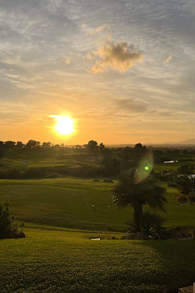 Ein weitläufiger, gepflegter Golfplatz mit Palmen und hügeligem Grün wird in warmes Licht eines Sonnenuntergangs getaucht, im Hintergrund Berge und weite Landschaft bei Palma de Mallorca.