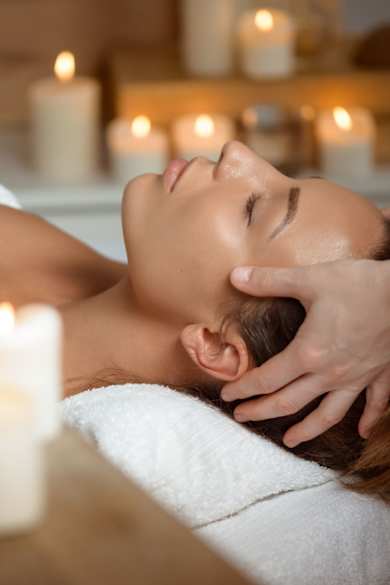 Eine Frau liegt mit geschlossenen Augen auf einer Massageliege und erhält eine entspannende Kopfmassage in stimmungsvoller Atmosphäre mit brennenden Kerzen