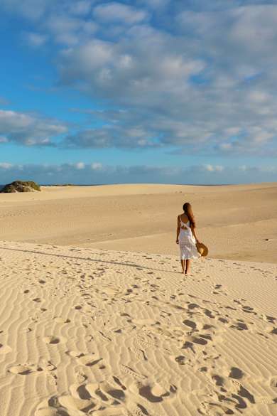 Eine Frau läuft im weißen Sommerkleid und mit Hut in der Hand über die Dünen bei Corralejo, Fuerteventura