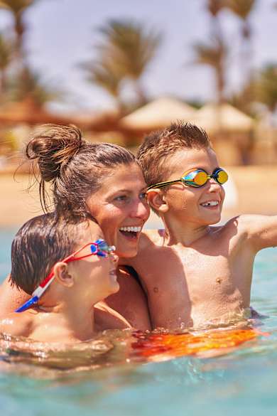 Eine Mutter ist mit ihren zwei Kindern im Wasser und hält sie im Arm. Im Hintergrund sieht man verschwommen den Strand und Palmen