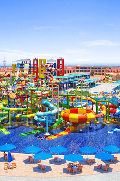 Luftaufnahme eines Wasserparks mit grünen, gelben und roten Rutschen, großem Poolbereich und zahlreichen blauen Sonnenschirmen rund um das Becken.