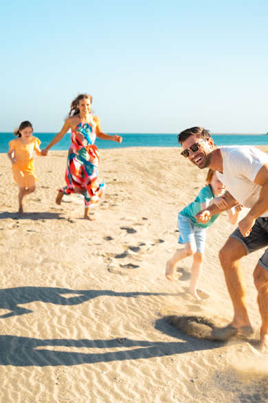 Eine Familie bestehend aus zwei Kindern, Mutter und Vater laufen spielerisch am Strand. Im Hintergrund ist das Meer zu sehen.