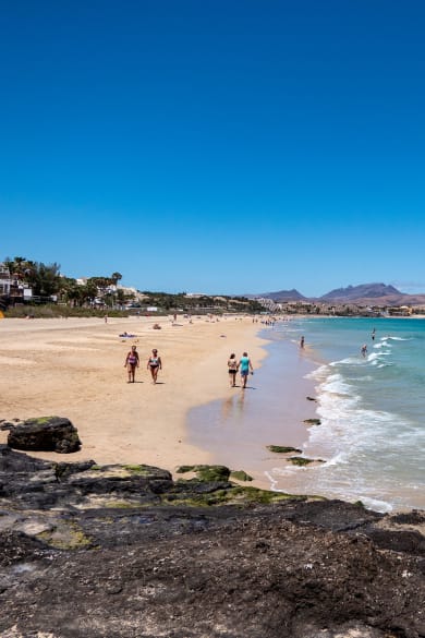 Langer Sandstrand mit Spaziergängern, klares blaues Meer, an der grünen Promenade reihen sich Häuser aneinander auf Fuerteventura