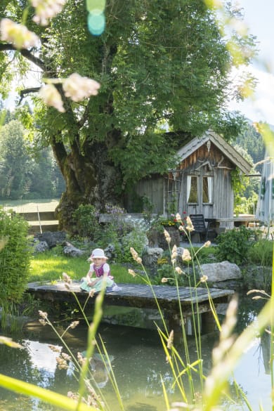 Ein Kind sitzt auf einem Steg an einem Teich. Im Hintergrund stehen ein großer Baum und eine Holzhütte.