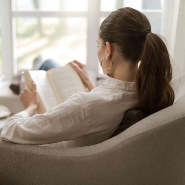 Blick von der Seite auf eine Frau, die mit hochgelegten Beinen in einem Sessel sitzt und ein Buch liest