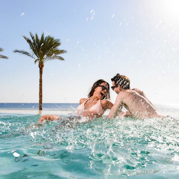 Ein Paar lacht im Pool mit Meer und einer Palme im Hintergrund.