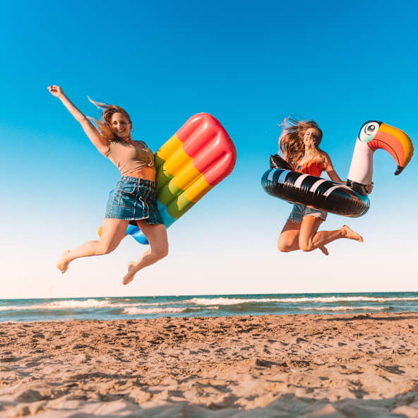 Drei Freundinnen springen mit aufblasbarem Spielzeug am Strand.