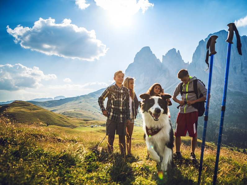 Familie steht mit Wanderausrüstung in den Dolomiten und Hund springt-durchs Bild © iStock.com/piola666