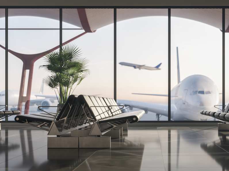 Innenansicht eines Flughafen-Terminals mit Sitzreihen und großer Fensterfront; draußen steht ein Flugzeug am Gate, während ein weiteres Flugzeug im Hintergrund startet.