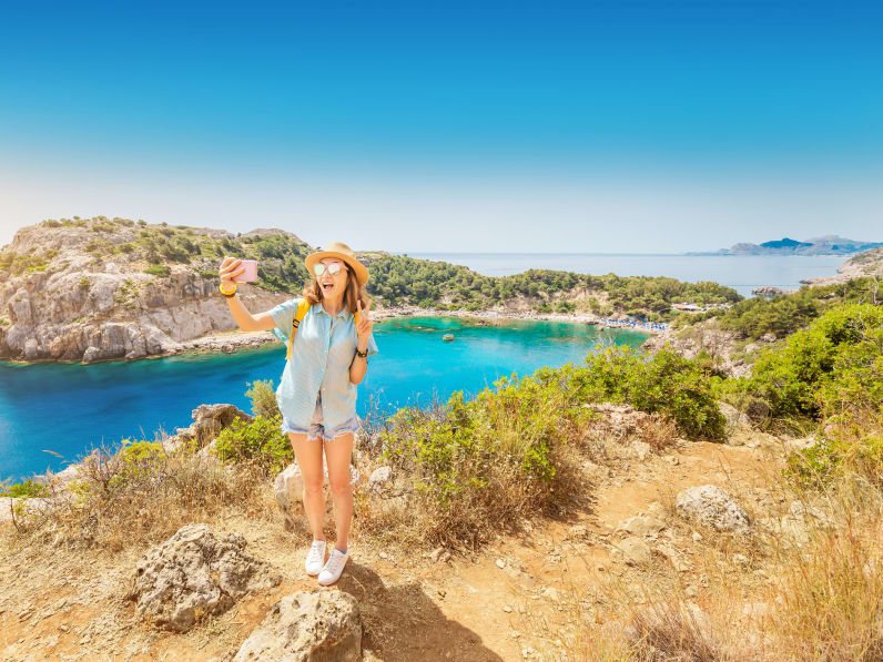 Mädchen mit Hut macht ein Selfie mit einer azurblauen Mittelmeer-Bucht im Hintergund © iStock.com/frantic00