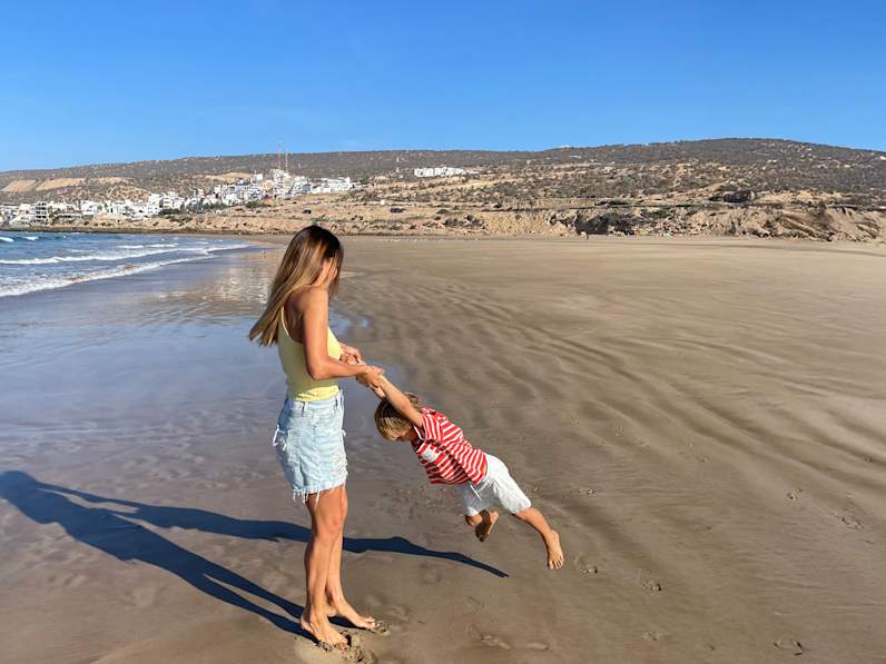 Eine Mutter spielt mit ihrem Sohn am Strand. Weit im Hintergrund erheben sich Hügel und ein Dorf.