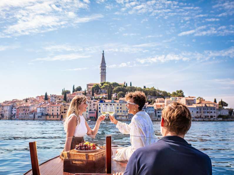 Zwei Frauen sitzen auf einem Boot auf dem Meer vor der Kulisse eines bunten Hafens in Kroatien und stoßen mit Weingläsern an