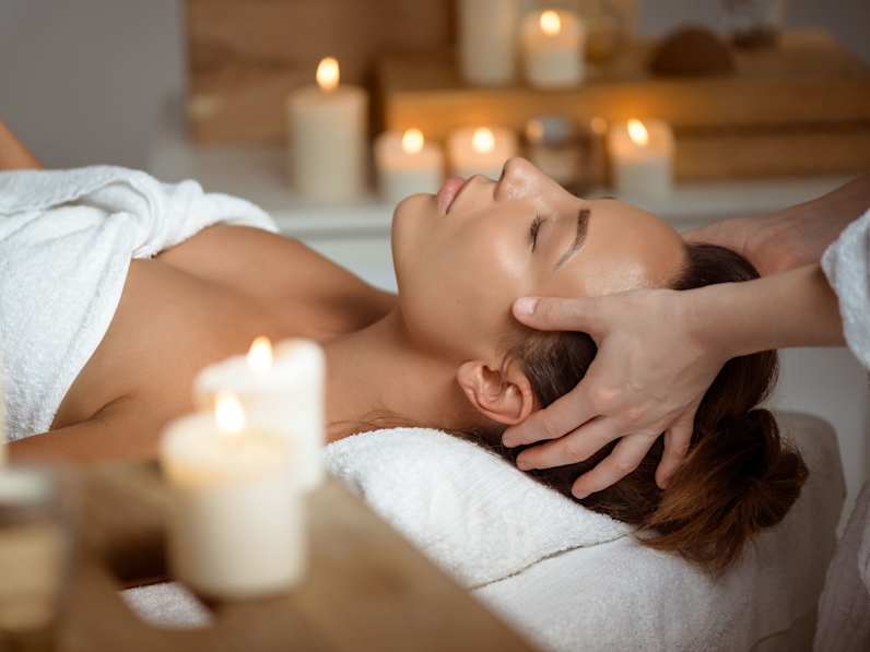 Eine Frau liegt mit geschlossenen Augen auf einer Massageliege und erhält eine entspannende Kopfmassage in stimmungsvoller Atmosphäre mit brennenden Kerzen