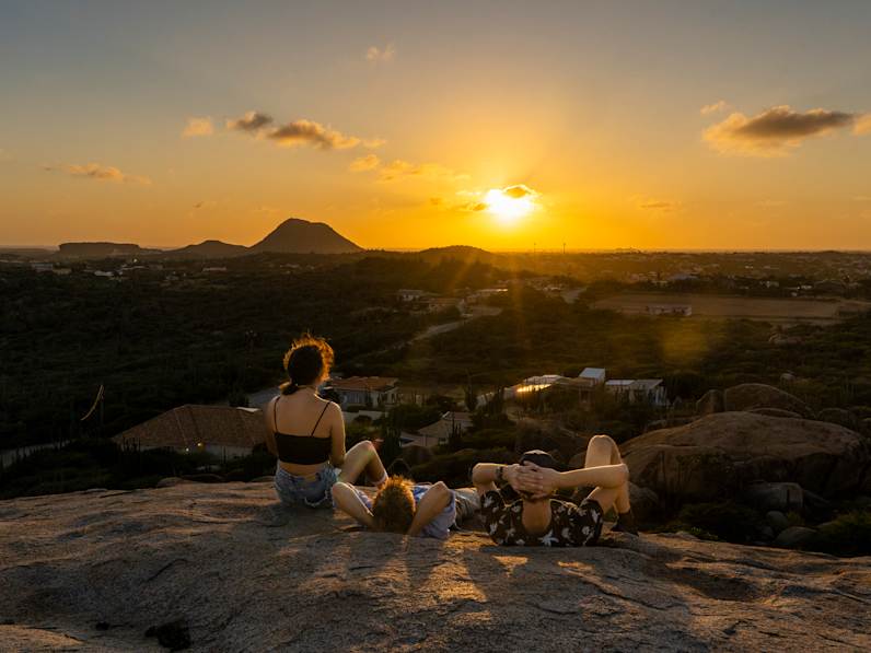 Drei junge Leute sitzen auf einem Felsen und schauen über eine Ebene auf den Sonnenuntergang.